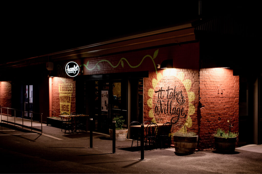 The entrance to Hamlet photographed at night, with the wall murals 'It takes a Village' and 'Things Happen Here' illuminated.